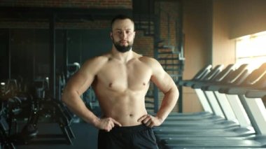Çıplak gövdeli, elleri belinde spor salonunda poz veren orta boy kaslı bir adam. Güçlü vücuda odaklan. Koşu bantları, arka planda geniş, karanlık bir spor salonu. Yüksek kalite 4k görüntü