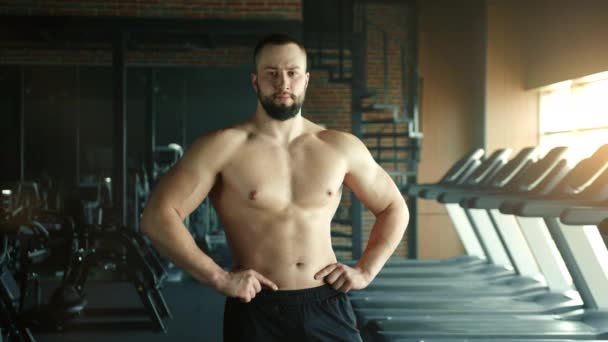 中等比例的肌肉男 赤裸的躯干 手放在腰部 在健身房摆姿势 专注于健康强壮的身体 跑步机 宽敞黑暗的健身房在后面 高质量的4K镜头 — 图库视频影像