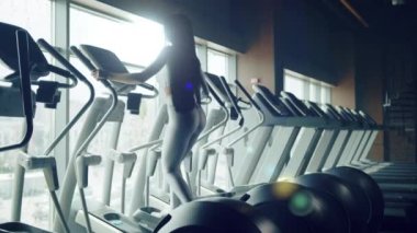 Siyah uzun saçlı, gümüş rengi spor giyen genç bir kadının eğitim süreci orbitrek çekiminde arkadan çekilir. Pencerelerdeki spor aletleri sırası. Yüksek kalite 4k görüntü