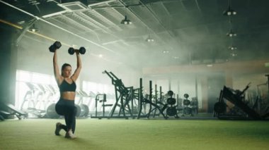 Formda genç bir kadın tarafından kolları başının üstüne kaldırılmış, spor bluzu, taytı ve spor ayakkabıları giymiş bir şekilde ileri doğru salınır. Arka planda tam teçhizatlı spor salonu. Yüksek kalite 4k görüntü