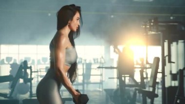 Spor salonunda dambıl gibi kıvrılan orta boy atletik kadın profili. Ağır çekim. Arka planda güneş ışınları olan tam teçhizatlı Misty spor salonu. Yüksek kalite 4k görüntü
