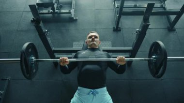 Spor salonunda göğüs presi yapan orta boylu yakışıklı bir genç. Adam siyah uzun kollu sıkıştırma gömleği ve mavi şort giyiyor. Yüz kasları çalışıyor. Yüksek kalite 4k görüntü