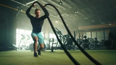 Sağlam vücutlu genç yakışıklı bir adam tarafından savaş halatında parçalanmış amcık dalgaları yapma süreci. Savaş halatları egzersizi. Arka planda tam teçhizatlı spor salonu. Yüksek kalite 4k görüntü