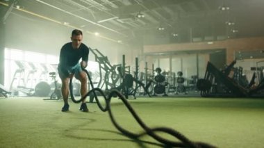 Üst kesimler, spor salonunda atletizm sporcusu tarafından yapılan savaş halatlarıyla birleşiyor. Hava bisikletleri, koşu bantları, kürek makinesi, arka planda sakallar. Yüksek kalite 4k görüntü