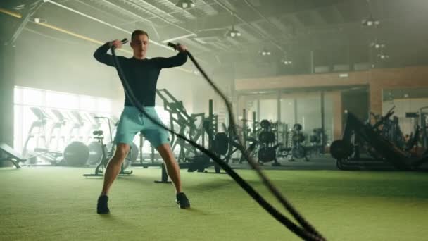 体操の戦闘ロープでジャッキ波をジャンプしているハンサムな運動能力の若い男 背景にはトレッドミル エアバイク ローイングマシン バーベル 高品質4K映像 — ストック動画