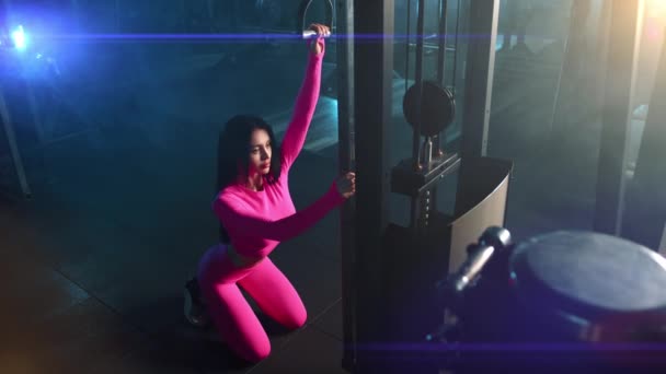 トレーナーケーブルマシン上で練習を行う運動若い美しい女性の高角度 女性は暗い照明のジムで膝の上にあります 謎の背景 高品質4K映像 — ストック動画