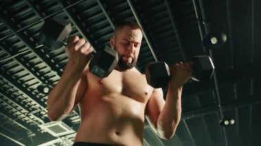 Çıplak ve terli gövdesi olan genç erkek, dambıllarla egzersiz yapıyor. Adam dambıl bisepsi kıvırmaya odaklanmış. Arka planda koyu tavan. Yüksek kalite 4k görüntü