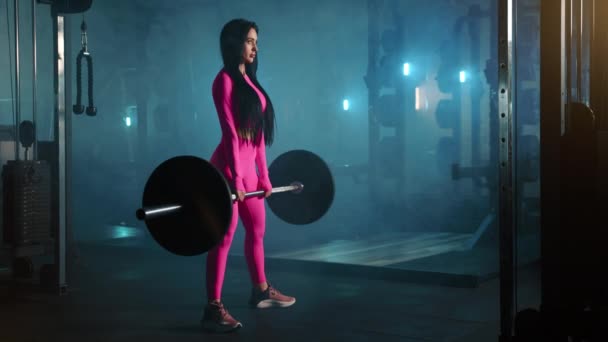 穿着粉色紧身运动服的女运动员在昏暗的灯光下在健身房做杠铃升降的全照 后备力量装备 高质量的4K镜头 — 图库视频影像