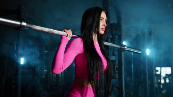 ピンクのスポーツトップでフィットボディの魅力的な女性のミディアムクローズアップバックスクワットを実行します サイドビュー 暗い照明ジム 謎の背景 高品質4K映像 — ストック動画