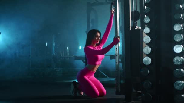 身穿粉色紧身运动上衣和腿的年轻而有运动魅力的女子在昏暗灯光下的健身房的教练缆机上做运动 模糊的背景 高质量的4K镜头 — 图库视频影像