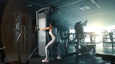 Formda kadın, pedin üzerinde otururken göğüs sineği makinesine bakar ve tam teçhizatlı spor salonunda egzersiz yapar. Gümüş rengi dar üst ve taytlar şekilleri vurguluyor. Yüksek kalite 4k görüntü