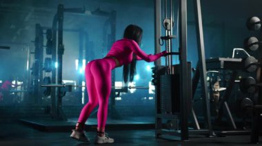 Pembe, dar spor kıyafetleri içindeki atletik kadın, koyu renk ışıklı spor salonundaki spor aletleriyle antrenör kablosu makinesinde glüt geri tepme gösterisi yapıyor. Yan görüş. Yüksek kalite 4k görüntü