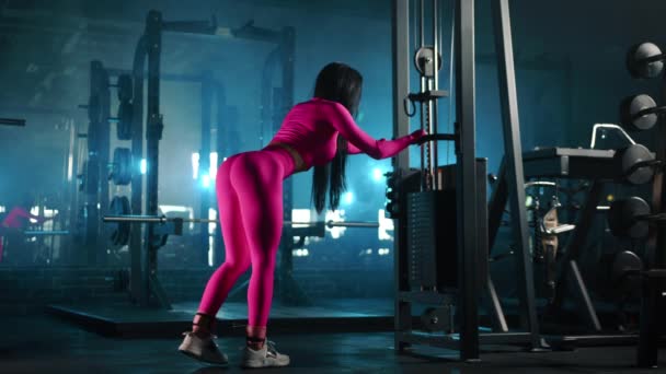 ピンクのスキンタイトなスポーツウェアのアスレチック女性は 強力な機器を備えた暗い照明ジムでトレーナーケーブルマシンで光学キックバックを実行します サイドビュー 高品質4K映像 — ストック動画