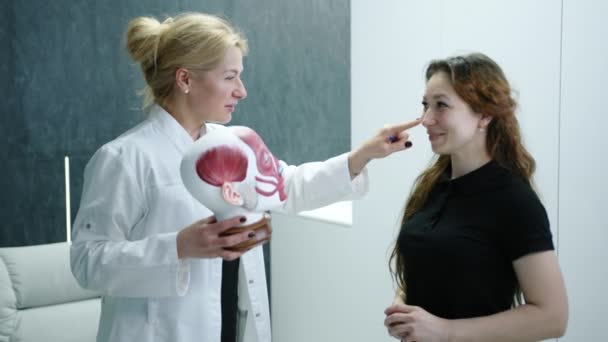 ブロンドの医者は女性患者に整形手術に関する相談を行う 専門家は頭部解剖モデルを使用する 患者は注意深く耳を傾け笑顔 高品質4K映像 — ストック動画