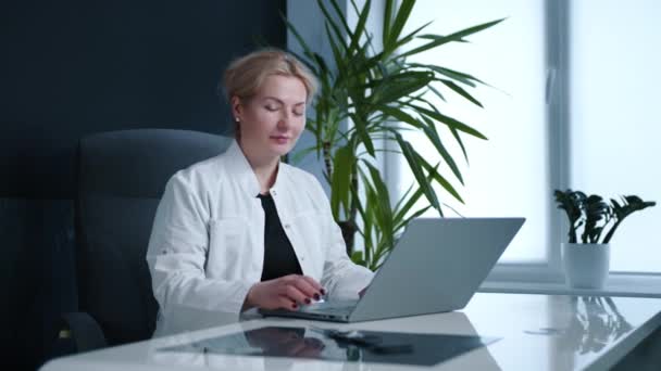 ブロンドの魅力的な医師のラップトップで入力し カメラで笑みを浮かべて 現代のオフィスに座っている ソフトオフィスチェア 白い机 背景に屋内ヤシの木 高品質4K映像 — ストック動画