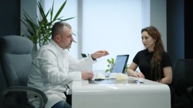 Orta yaşlı erkek doktor, göğüs implantı olan kadın hastaya danışmanlık yapıyor. Dolabın içinde modern bir tasarım ve iç palmiye ağacı var. Silikonların modelleri. Yüksek kalite 4k görüntü