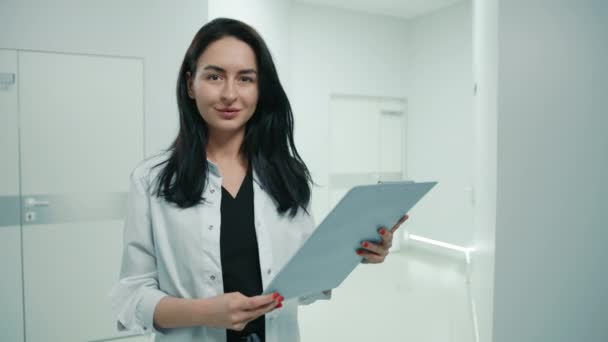 現代の診療所のファイルフォルダと研究室のコートでかわいい若い暗い髪の女性医師 医者はカメラと笑顔を見ている 背景に白い回廊 高品質4K映像 — ストック動画