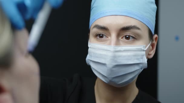 医療用マスク 帽子や手袋の女性医師の肩のショットの上に化粧品の治療のための女性の顔の皮膚マーカーペンで外科的マーキングを作ります 高品質4K映像 — ストック動画
