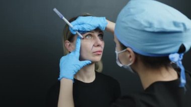 Kadın yüzünün cerrahi müdahalesi sırasında omzundan vurulmuş. Kadın doktor göz altı işaretleme yapıyor, burun kıvrımları deri kalem kullanıyor. Yüksek kalite 4k görüntü