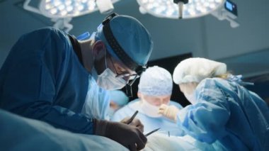 Genç erkek cerrah estetik ameliyat sırasında hastayı dikiyor. Doktor dikiş atar. Arka planda cerrahi ekip var. Operasyon ışığı öncülüğünde. Yüksek kalite 4k görüntü