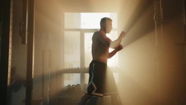 太陽の光で照らされたジムでシャドウボクシングのトレーニングを行う裸の筋肉質の胴を持つ若いフィット男 プロファイルショット 背景の窓の近くにあるダンベルラック 高品質4K映像 — ストック動画
