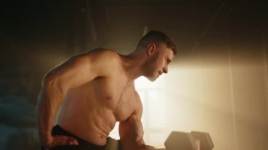 Profilde genç bir sporcunun güneş ışığında oturup dambılını kaldırdığı görülüyor. Bir elini beline koy. Sporcu pazularına bakıyor. Yüz kasları gergin. Yüksek kalite 4k görüntü