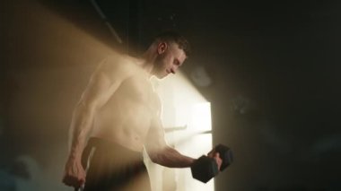 Tek kollu dambıl bisepsi yapan genç bir sporcunun profil fotoğrafı. Güneş ışığında spor salonunda duruyor. Adam bir elini daha beline koyar. Yüz kasları gergin. Yüksek kalite 4k görüntü