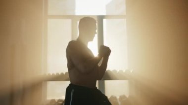 Spor salonunda güneş ışınlarıyla boks yapan tek başına bir boksör. Çıplak kaslı sporcu hava yumrukları atıyor. Pencerede aptal askısı var. Yüksek kalite 4k görüntü