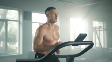 Koşu bandında yavaş koşan genç bir sporcunun orta boy fotoğrafı. Sonra da spor salonunda koşuyor. Çıplak gövde kaslı ve terli. Eller ileri geri hareket ediyor. Yüksek kalite 4k görüntü