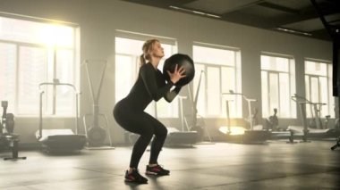 Siyah dar spor giyimli, formda genç bir kadının tıbbi topu elinde tutarak çömeldiği profil fotoğrafı. Kadınlar hafif ferah spor salonunda egzersiz yapıyor. Yüksek kalite 4k görüntü