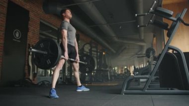 Genç yakışıklı atletik adam geniş spor salonunda halteri kaldırıyor ve siyah renkli güçlü eğitim ekipmanlarıyla. Sporcu kaldırma hareketi yapıyor, bacaklarını ayırıyor. Yüksek kalite 4k görüntü
