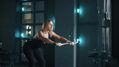 Yakışıklı sarışın, kablolu yayın makinesinde egzersiz yapıyor. Aydınlatılmış karanlık spor salonunda kadın kablolu yayın yapıyor. Kadın aktif olarak egzersiz yapıyor. Yüksek kalite 4k görüntü