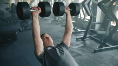 Genç bir sporcunun spor salonunda ağırlık kaldırırken orta boy fotoğrafı. Adam bankta yatar, kollarını dikey olarak uzatır ve dambılları indirir. Yüksek kalite 4k görüntü