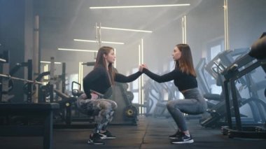 Yan yana duran, el pençe divan duran, el ele çömelmiş iki genç kadının profili. Arka planda tam teçhizatlı spor salonu var. Yüksek kalite 4k görüntü