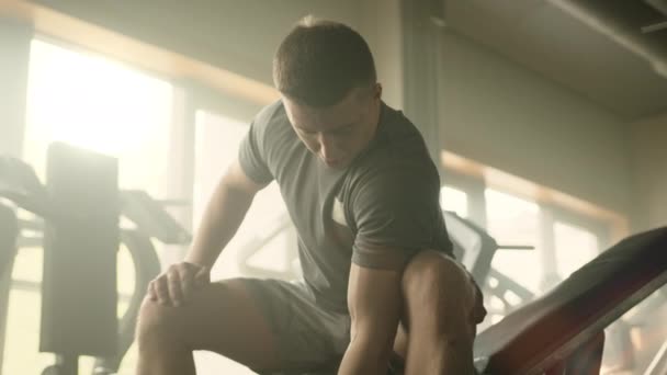 ベンチに座っている若い選手によってカール片腕の両足を実行するプロセス 男はビキップを見てる 軽い設備の整ったジムでスポーツマンの練習 高品質4K映像 — ストック動画