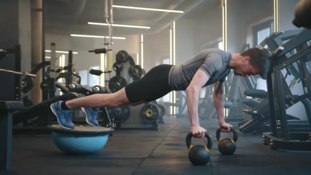 年轻健康的男人在健身房用水壶和平衡板做俯卧撑 水平位置的运动员拿着两只水壶 脚踏在平衡板上 高质量的4K镜头 — 图库视频影像
