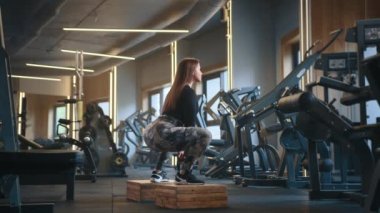 Atletik genç, uzun saçlı bir kadının tahta bloklara çömelmiş bir şekilde çömeldiği profil fotoğrafı. Ağır çekim. Arka planda tam teçhizatlı spor salonu. Yüksek kalite 4k görüntü