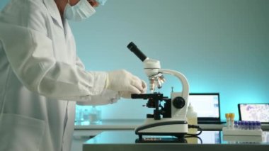 Tıbbi eldivenleri, maskesi, şapkası ve cüppesi olan orta yaşlı bir erkek laboratuvar çalışanı laboratuvar araştırmaları için mikroskoba cam parçası yerleştiriyor. Laptop, tablet, test tüpleri. Yüksek kalite 4k görüntü