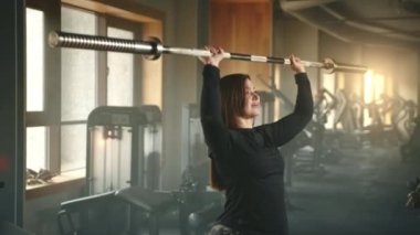 Spor salonunda Olimpik halterle tepenin üstünde dans eden tatlı bir kadının orta boy fotoğrafı. Kadın halteri yukarı kaldırıyor ve birkaç saniye tutuyor. Yüksek kalite 4k görüntü