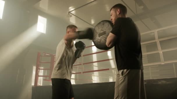 ボクシングリング近くのジムで中年フィットボクサーによってボクシングパッドでパンチを投げるプロセス 他のスポーツマンはボクシングパッドを保持しながらボクサー演習 高品質4K映像 — ストック動画
