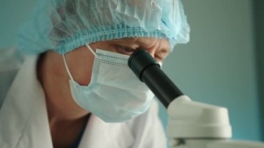 Medikal maskeli, şapkalı ve laboratuvar önlüklü orta yaşlı bir doktor mikroskoba bakıyor. Tıp çalışanı laboratuvar araştırmalarına odaklanmış durumda. Yüksek kalite 4k görüntü