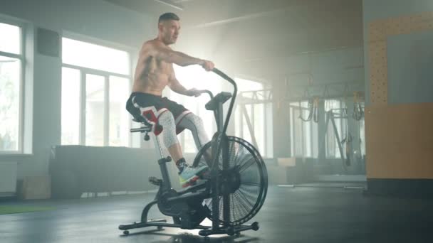エアバイクで汗びっしょりの素足の胴を持つ若いスポーツマンは 手足がフットレストに残りながら手仕事 それから男はペダルに足を置き ペダルを踏み始めます 高品質4K映像 — ストック動画