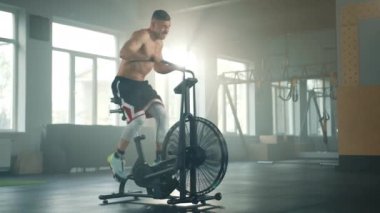 Geniş spor salonunda hava bisikleti üzerinde aktif egzersiz yapan çıplak kaslı genç bir sporcu. Sporcu kendini yorgun hissederek egzersiz yapmayı bırakır. Yüksek kalite 4k görüntü