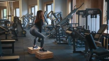 Atletik kadın, tahta blokların üzerinde, ayakları omuz genişliğinde, kettlebell ile çömelme hareketleri yapıyor. Dayanıklı eğitim ekipmanlarıyla donatılmış spor salonu. Yüksek kalite 4k görüntü