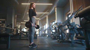 Siyah spor tişörtlü, sağlıklı vücutlu, kahverengi saçlı ve gri tozlu genç bir kadın tam teçhizatlı spor salonunda halter kaldırıyor. Profil çekimi, yavaş çekim. Yüksek kalite 4k görüntü