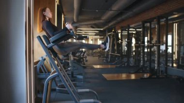 Tam teçhizatlı spor salonunda basın için dikey bacak yükseltme yapan genç bir kadın. Kadın düz bacaklar kaldırıyor, kolları yastıklı parmaklıklar üzerinde, vücudu düz. Yüksek kalite 4k görüntü