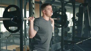 Tam teçhizatlı spor salonunda halter çökerken orta boy yakışıklı bir atlet. Adam halteri tuzaklarda ve omuzlarda tutuyor ve çömelme hareketleri yapıyor. Yüksek kalite 4k görüntü