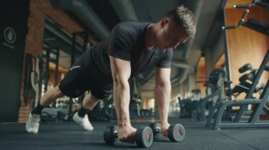 Genç sporcu, tam teçhizatlı spor salonunda halterlerle şınav çekiyor. Sporcu, şınav pozisyonunda dirsekler yukarı, göğüs zırhına ağırlık getiriyor. Yüksek kalite 4k görüntü