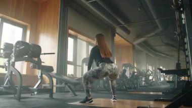 Amerikan kettlebell salıncaklarını seslendiren atletik genç bir kadın. Kadın tencere dibin kara, seninki benden kara. Arka planda güçlü eğitim ekipmanları var. Yüksek kalite 4k görüntü