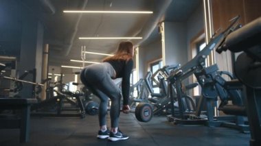 Formda kalçaları olan atletik genç bir kadının geniş spor salonunda halterle kaldırma performansı sergilemesi. Arka planda güçlü eğitim ekipmanları var. Yüksek kalite 4k görüntü